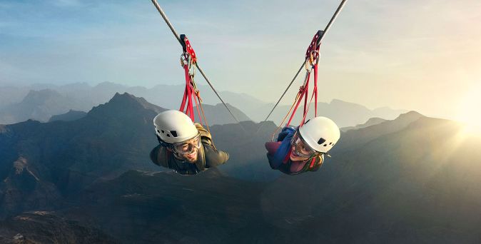 Zwei Personen mit weißen Helmen liegen in Gurten an einer Zipline über bergiger Landschaft bei Sonnenuntergang in Ras Al Khaimah.