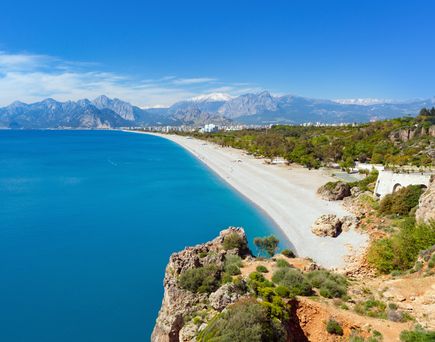 Panoramablick auf den Konyaalti-Strand in Antalya, Türkei, mit türkisblauem Meer, breitem Kiesstrand und grünen Parkanlagen vor der Kulisse des Taurusgebirges unter klarem, blauem Himmel
