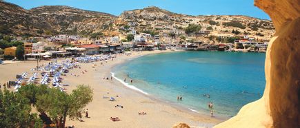 Blick von einer Felsöffnung auf einen Sandstrand mit mehreren Sonnenschirmen, Badegästen und einem Dorf am Hang.