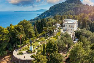 Luftaufnahme des Achilleion-Palasts mit umliegendem Garten, Palmen und Blick auf das Meer bei klarem Himmel.
