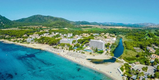 Luftaufnahme eines Hotels und Strandes mit Sonnenschirmen am türkisfarbenen Meer, umgeben von grünen Hügeln und einem Fluss