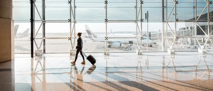 Frau mit Rollkoffer läuft an einer großen Glaswand entlang durch ein modernes Flughafenterminal, während im Hintergrund Flugzeuge auf dem Rollfeld stehen