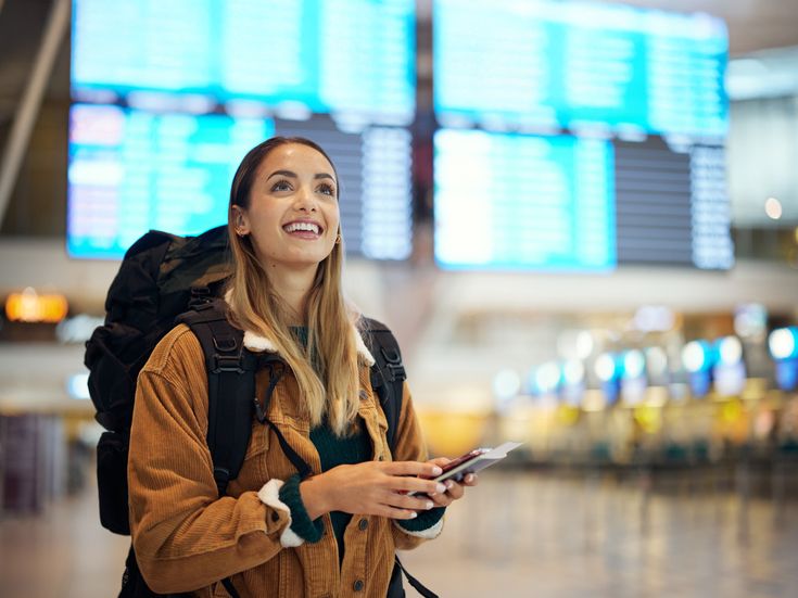 Reisende Frau mit Rucksack steht lächelnd am Flughafen vor einer großen Anzeigetafel und hält Reisedokumente in der Hand