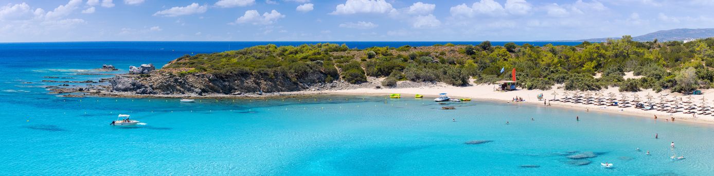 Strand mit Reihen von Sonnenschirmen und Liegen, türkisfarbenes Wasser mit Schwimmern, bewaldete Küste und Hügel im Hintergrund unter blauem Himmel mit Wolken.