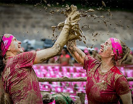 Zwei Frauen mit schlammverschmierter Kleidung und pinken Bändern schlagen zusammen ein und der Schlamm spritzt während sie den Muddy Angel Run machen