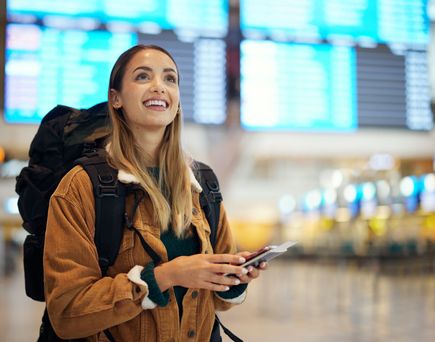 Junge Frau mit Rucksack, Smartphone und Reisepass lächelt in einer Flughafenhalle vor den großen Anzeigetafeln