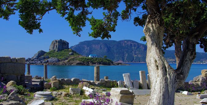 Ruinen mit Säulen am Ufer vor einer kleinen bewachsenen Insel und Bergen im Hintergrund unter blauem Himmel