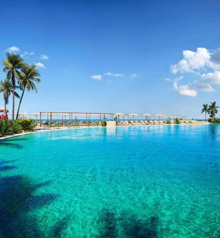 Großer Außenpool mit klarem Wasser, umgeben von Liegestühlen und Sonnenschirmen, blauer Himmel mit wenigen Wolken.