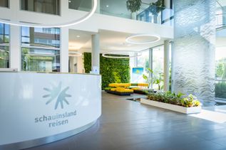 Moderner Empfangsbereich im Gebäude 3 von schauinsland-reisen mit weißem Empfangstresen und Firmenlogo, großzügigem Luftraum, ringförmigen Deckenleuchten, Pflanzen und Sitzbereich im Hintergrund