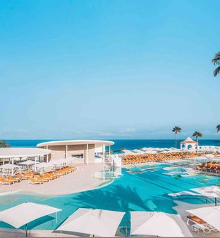 Großer Hotelpool mit mehreren weißen Sonnenschirmen und orangefarbenen Liegestühlen, Palmen und Meer im Hintergrund unter blauem Himmel.