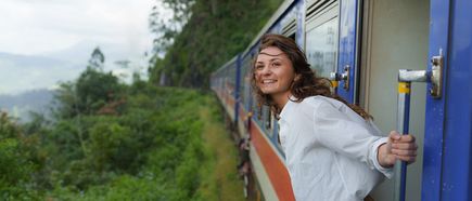 Frau lehnt sich aus der geöffneten Tür eines fahrenden Zuges und blickt auf eine grüne, hügelige Landschaft
