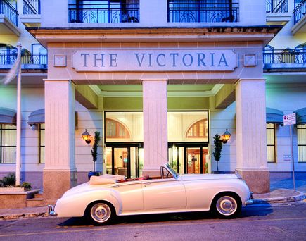 Klassischer weißer Cabrio vor dem Eingangsbereich des Hotels "The Victoria" mit Säulen und großen Fenstern.