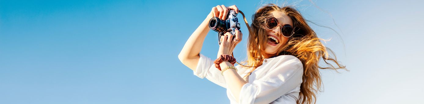 Frau mit langen, wehenden Haaren hält eine Kamera über dem Kopf, trägt weiße Bluse und Armbänder, blauer Himmel im Hintergrund.