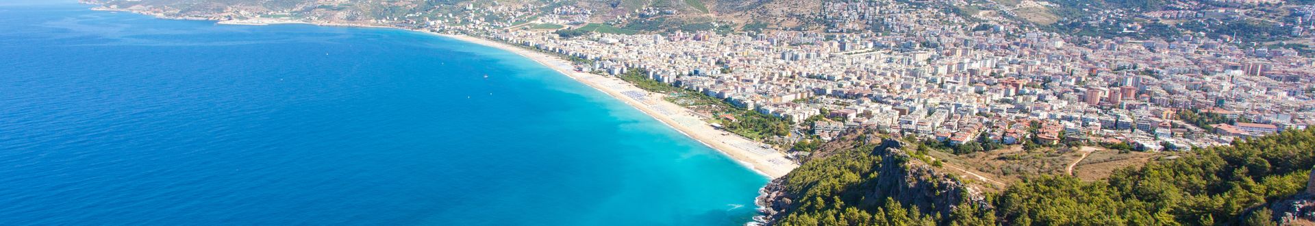 Panoramablick auf den Kleopatra-Strand in Alanya, Türkei, mit türkisblauem Meer, feinem Sandstrand und der dahinterliegenden Stadt, umgeben von grünen Hügeln