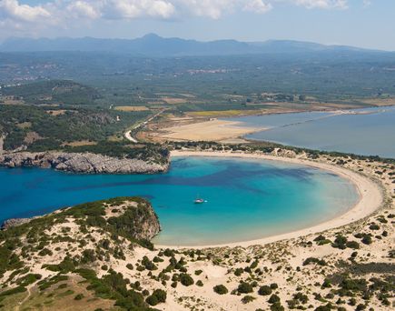 Luftaufnahme einer geschwungenen Sandbucht mit klarem blauem Wasser und bewachsenen Dünen im Vordergrund.