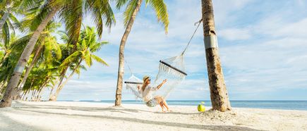 Person sitzt in einer Hängematte zwischen Palmen an einem weißen tropischen Sandstrand mit Blick auf das ruhige Meer