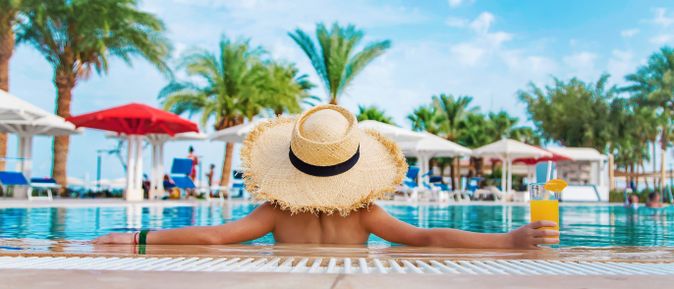 Person mit großem Strohhut lehnt an der Kante eines Pools und hält ein Getränk, während Palmen, Sonnenschirme und Liegen eine tropische Hotelanlage umgeben