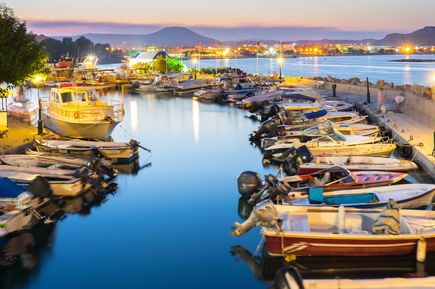 Kleiner Hafen mit mehreren Booten am Ufer, beleuchtete Stadt und Berge im Hintergrund bei Abenddämmerung.