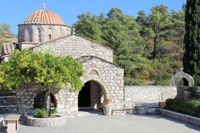 Steinernes Klostergebäude mit Rundkuppel und Kreuz, umgeben von Bäumen und einer gepflasterten Fläche mit Sitzbank und Pflanzenkübeln.