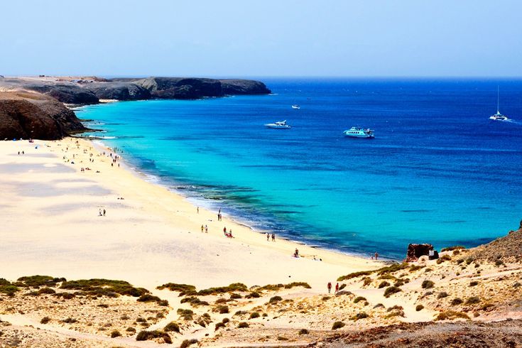 Strand mit hellem Sand, dahinter türkisblaues Meer und mehrere Boote, im Vordergrund felsige Küste