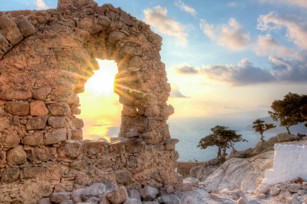 Sonnenstrahlen scheinen durch ein rundes Fenster in einer alten Steinmauer mit Blick auf das Meer und Bäume am felsigen Ufer.