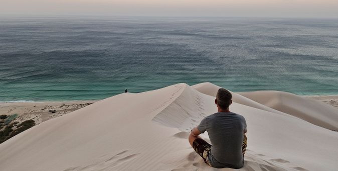 Ein Mann sitzt auf einer Sanddüne im Jemen, Socotra und blickt aufs tiefblaue Meer hinaus bis zum Horizont, welcher im Himmel verschwindet.