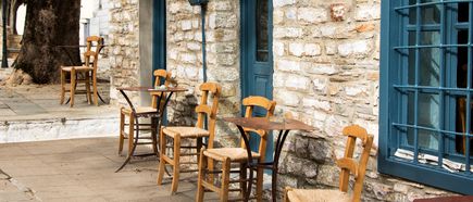 Mehrere Holzstühle mit geflochtenen Sitzen stehen vor einem steinernen Gebäude mit blauen Türen und Fenstern.