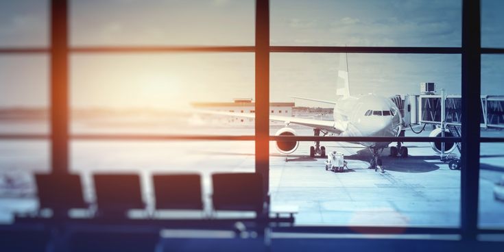 Blick durch die Fensterfront eines Flughafenterminals auf ein angedocktes Passagierflugzeug am Gate, mit Sitzreihen im Vordergrund und sanftem Morgen- oder Abendlicht.