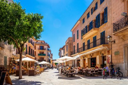 Straßencafé in der Altstadt von Alcudia auf Mallorca mit historischen Häusern, Sonnenschirmen und Gästen unter blauem Himmel