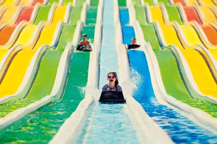Mehrere Kinder rutschen auf bunten, parallel verlaufenden Wasserrutschen in einem Wasserpark auf Matten hinab