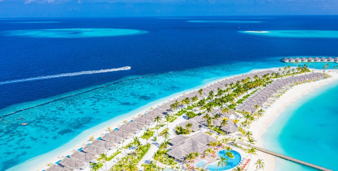 Luftaufnahme eines Resorts auf den Malediven mit mehreren Bungalows, umgeben von türkisfarbenem Wasser und weißen Sandstränden. Im Hintergrund sind kleine Bootsspuren im Wasser sichtbar.