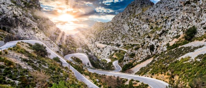 Serpentinenstraße in der Serra de Tramuntana auf Mallorca, die sich durch steile Felslandschaft windet, mit Sonnenuntergang im Hintergrund