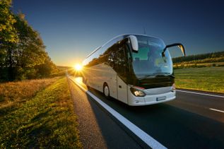 Moderner Reisebus fährt bei Sonnenuntergang über eine Landstraße, umgeben von Feldern und Bäumen