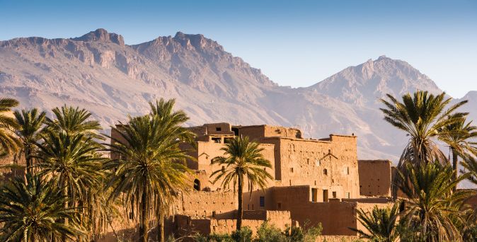 Lehmgebäude umgeben von Palmen vor einer Berglandschaft unter klarem Himmel in Marokko