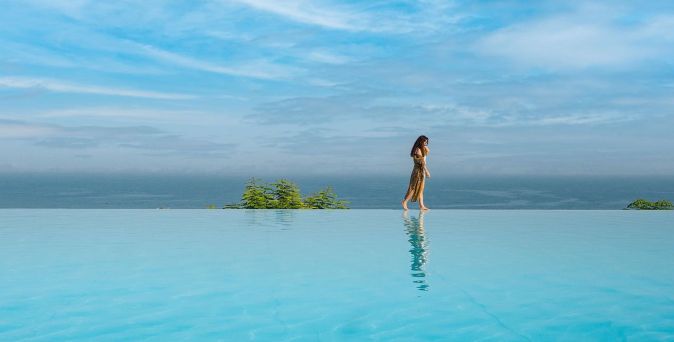 Frau läuft am Rand eines Infinity-Pools mit Blick auf den blauen Himmel und das Meer 