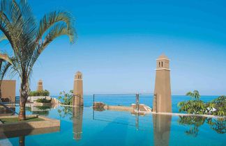 Zwei braune Türme neben einem Infinity-Pool mit Blick auf das Meer und einen klaren blauen Himmel, links eine Palme.