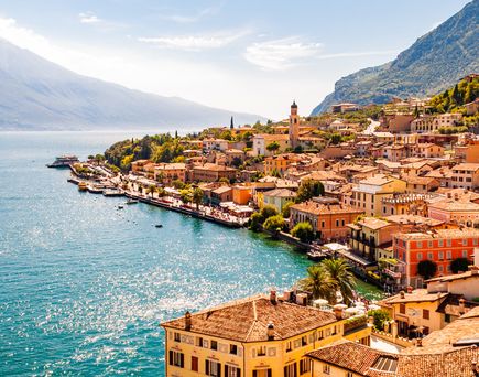 Blick auf eine belebte Uferstadt am Gardasee in Norditalien, umgeben von Bergen und klarem Wasser