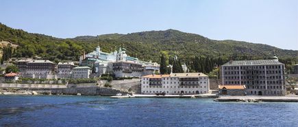 Blick auf mehrere Klostergebäude am Ufer vor bewaldeten Hügeln unter klarem Himmel.