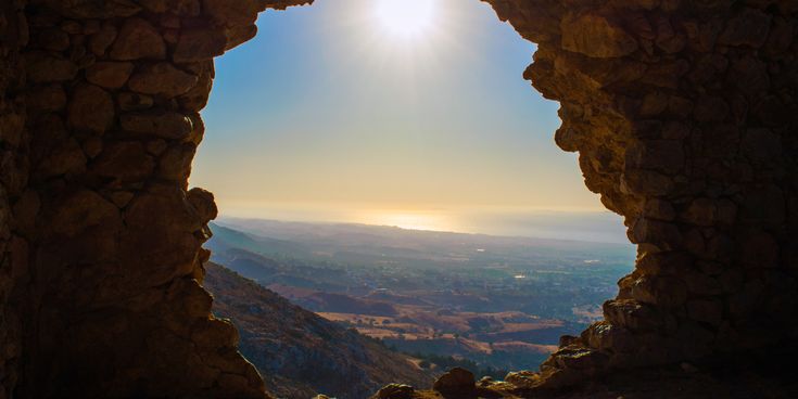 Blick durch eine Felsöffnung auf eine weite Landschaft mit Hügeln und einem hellen Himmel mit Sonne