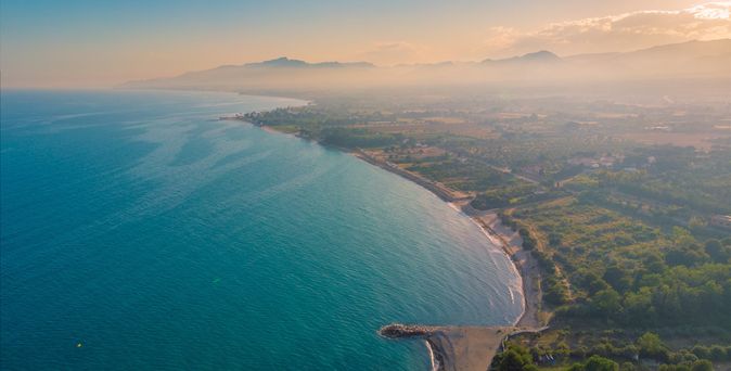 Drohnen-Luftbild mit Blick auf die Küste der Costa Dorada, Katalonien, Spanien, endlos lange Buchten mit begrüntem Hinterland bei Sonnenaufgang, tiefblaues Meer glitzert vom Sonnenlicht, leicht nebelig am Horizont