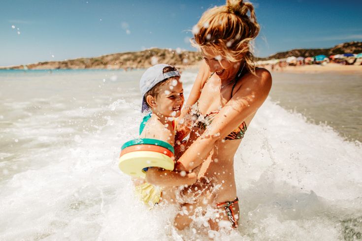 Mutter spielt lachend mit ihrem kleinen Sohn im Meer, während Wellen spritzen