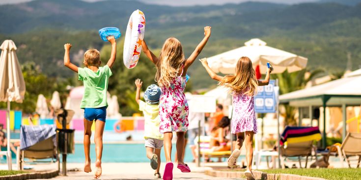 Mehrere Kinder laufen auf einer Hotelanlage in Richtung Poolbereich, umgeben von Liegen, Sonnenschirmen und einer Bergkulisse im Hintergrund