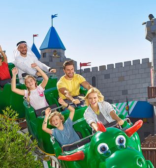 Grüne Drachenachterbahn mit mehreren Fahrgästen, im Hintergrund eine Burg mit blauen Türmen und Fahnen im Legoland Dubai.