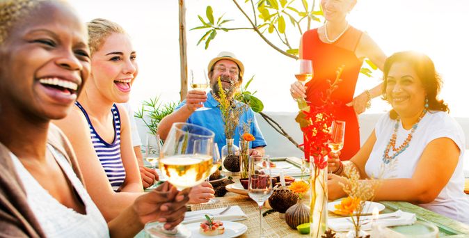 Fünf Personen sitzen an einem gedeckten Tisch mit Gläsern und Speisen, eine Person steht und hält ein Glas, Tisch mit Kerzen und Dekoration.