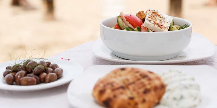Weißer Teller mit griechischem Salat, daneben Teller mit Oliven und ein Teller mit Fladenbrot und Kräuterdip auf weißem Tischtuch.