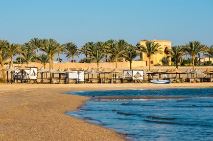 Strand von Coraya Bay in Ägypten mit Palmen, Sonnenschirmen, weißen Strandliegen und ruhigem Meer