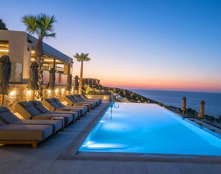 Beleuchteter Infinity-Pool mit Liegen und Palmen vor einem Hotel, Blick aufs Meer bei Sonnenuntergang.