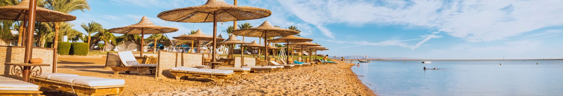 Panoramablick auf den Sandstrand am Roten Meer in Ägypten mit Strohdach-Sonnenschirmen, Liegen und Palmen, ruhiges Meer und blauer Himmel mit Wolken
