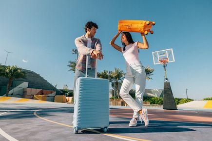 Zwei Personen mit American Tourister Koffern auf einem Basketballplatz unter blauem Himmel. Eine Person hält einen orangefarbenen Koffer über dem Kopf, während daneben ein hellblauer Rollkoffer steht.