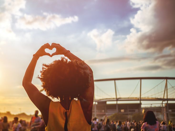Person formt mit den Händen ein Herz vor dem Abendhimmel auf einem Festivalgelände, während sich im Hintergrund Besucher und eine Veranstaltungsbühne im warmen Licht der Abendsonne befinden